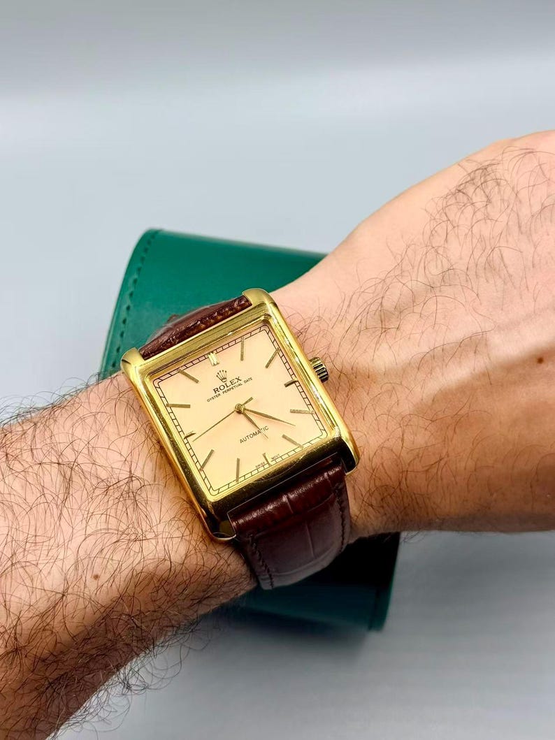 May include: A gold-toned rectangular Rolex Oyster Perpetual Date automatic watch with a light-colored face and brown leather band. The watch has a gold bezel and a brown leather strap. The watch is on a wrist.