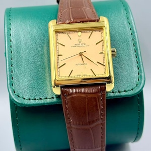May include: A gold-toned Rolex Oyster Perpetual Date automatic watch with a rectangular face and brown leather band. The watch face is a light peach color with black hands and markers. The watch is displayed on a green leather case.