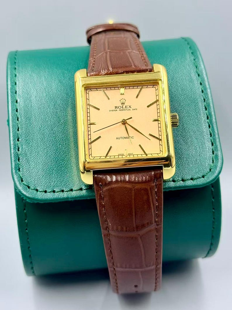 May include: A gold-toned Rolex Oyster Perpetual Date automatic watch with a rectangular face and brown leather band. The watch face is a light peach color with black hands and markers. The watch is displayed on a green leather case.