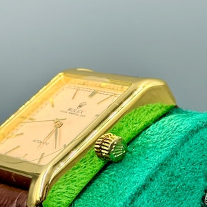 May include: A gold-toned Rolex wristwatch with a rectangular face and a brown leather band. The watch face is a light peach color with gold hands and markers. The watch is set against a green textured background.