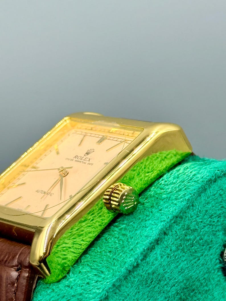May include: A gold-toned Rolex wristwatch with a rectangular face and a brown leather band. The watch face is a light peach color with gold hands and markers. The watch is set against a green textured background.