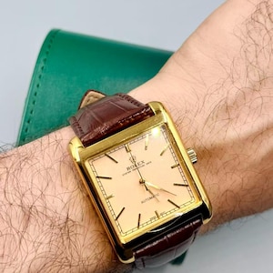 May include: A gold-toned rectangular wristwatch with a light peach-colored face and brown leather band. The watch face displays the text "ROLEX OYSTER PERPETUAL DATE AUTOMATIC". The watch is on a wrist.