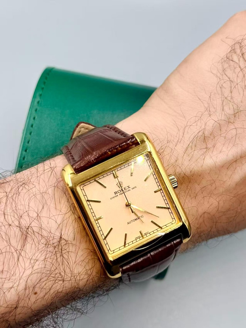 May include: A gold-toned rectangular wristwatch with a light peach-colored face and brown leather band. The watch face displays the text "ROLEX OYSTER PERPETUAL DATE AUTOMATIC". The watch is on a wrist.