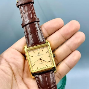 May include: A gold-toned rectangular Rolex watch with a light-colored face and black hands. The watch has a brown leather band with a crocodile pattern. The watch is an automatic model.