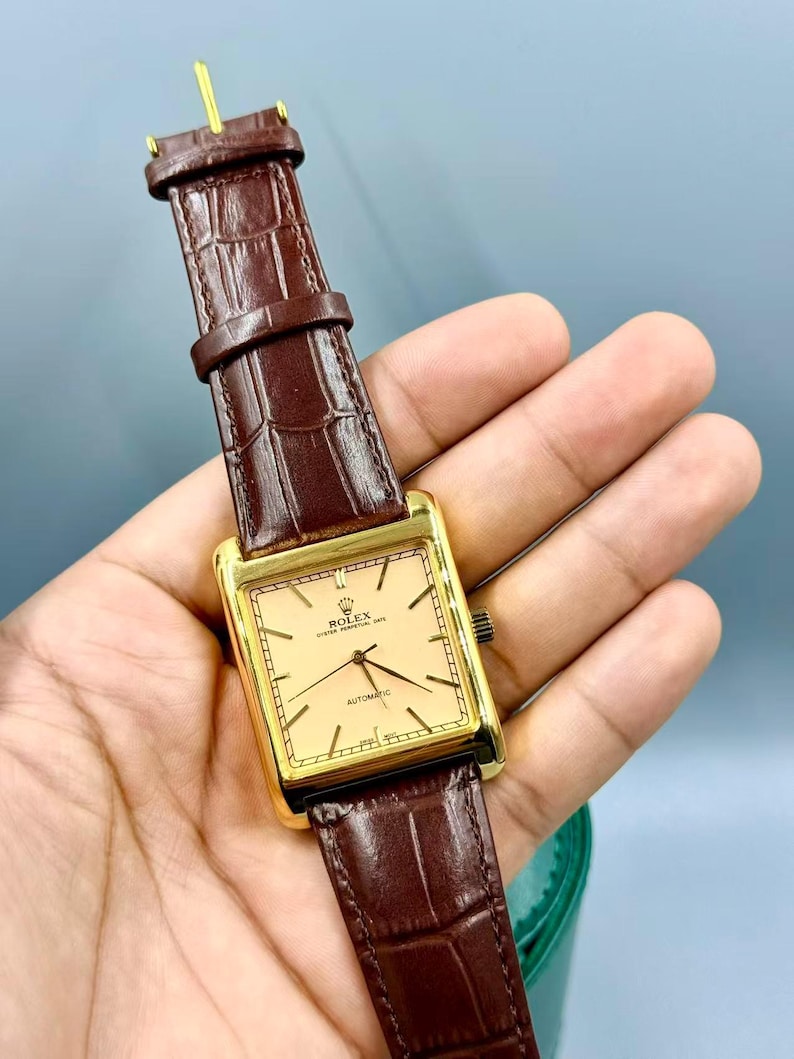 May include: A gold-toned rectangular Rolex watch with a light-colored face and black hands. The watch has a brown leather band with a crocodile pattern. The watch is an automatic model.