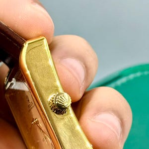 May include: Close-up of a gold-toned rectangular watch case with a textured crown. The watch has a brown leather strap. The watch face is visible, with the brand name "Rolex" printed on it.