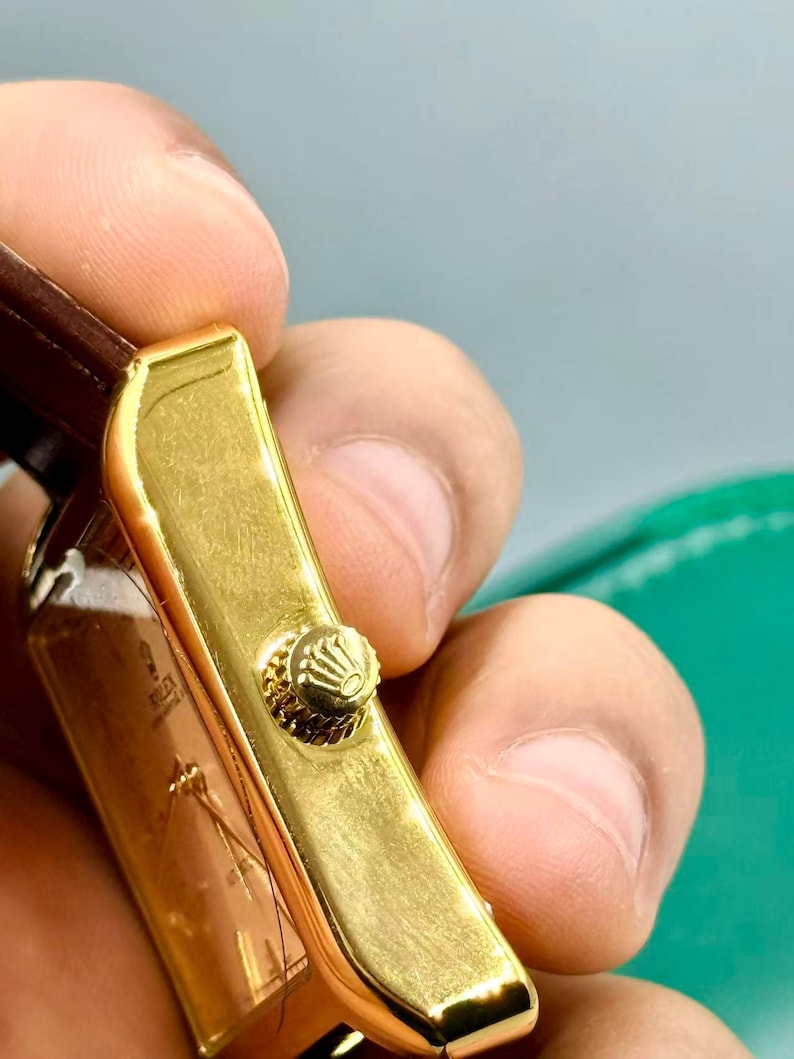 May include: Close-up of a gold-toned rectangular watch case with a textured crown. The watch has a brown leather strap. The watch face is visible, with the brand name "Rolex" printed on it.