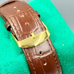 May include: Close-up of a brown leather watch strap with a gold-colored buckle. The strap has a textured, crocodile-like pattern and is stitched along the edges. The buckle is rectangular and shiny.