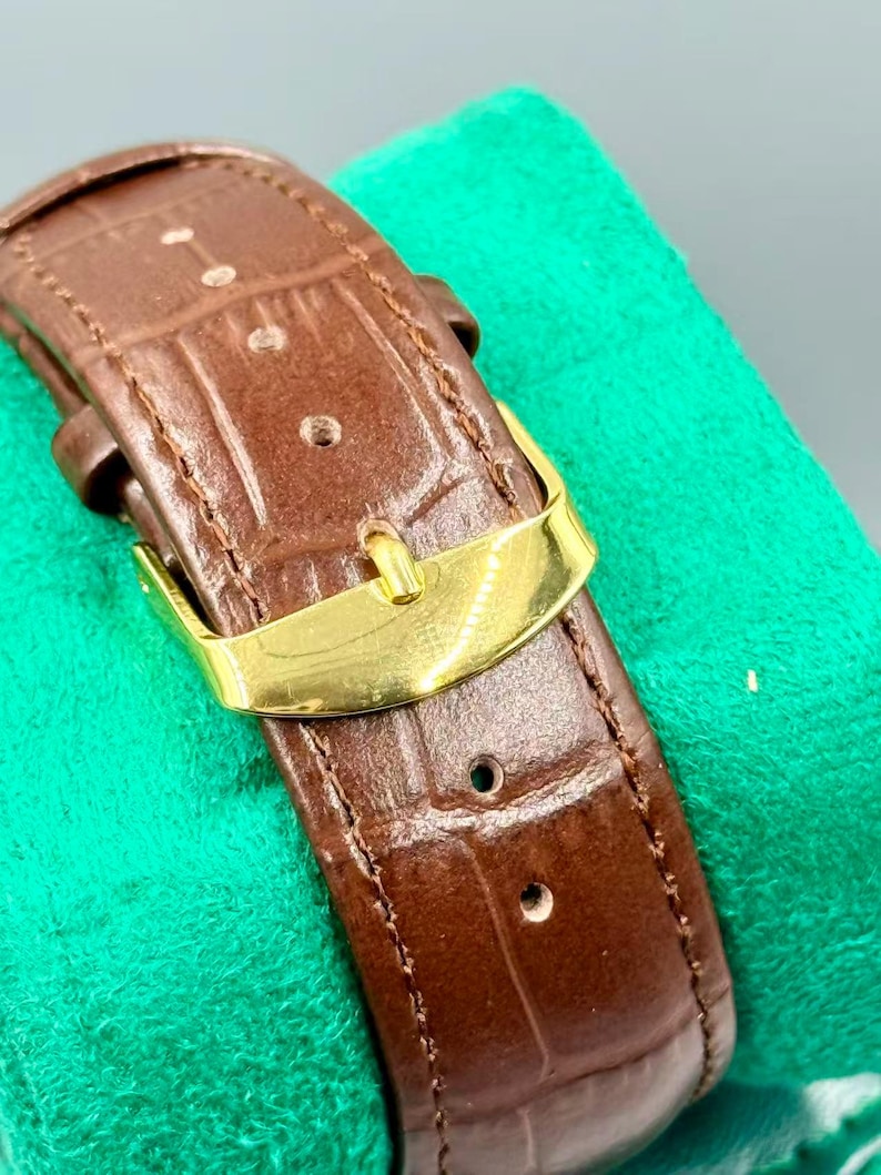 May include: Close-up of a brown leather watch strap with a gold-colored buckle. The strap has a textured, crocodile-like pattern and is stitched along the edges. The buckle is rectangular and shiny.