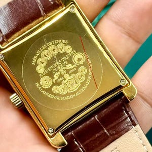 May include: Close-up of a gold rectangular watch case with a brown leather strap. The watch back features engraved text and a circular emblem. The strap is a dark brown color with a textured pattern.