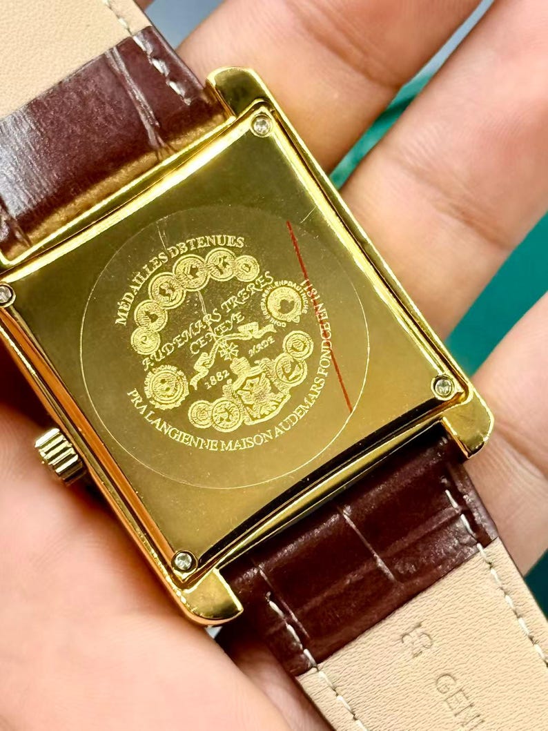 May include: Close-up of a gold rectangular watch case with a brown leather strap. The watch back features engraved text and a circular emblem. The strap is a dark brown color with a textured pattern.