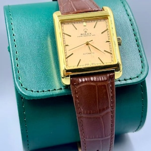 May include: A gold-toned Rolex Oyster Perpetual Date automatic watch with a rectangular face and brown leather band. The watch face is a light peach color with gold hands and markers. The watch is resting on a green leather case.