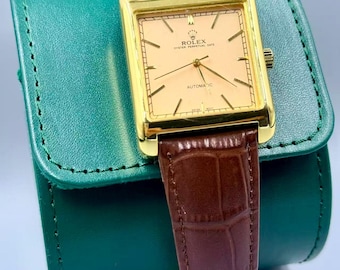 Può includere: Un orologio automatico Rolex Oyster Perpetual Date color oro con quadrante rettangolare e cinturino in pelle marrone. Il quadrante dell'orologio è color pesca chiaro con lancette e indicatori dorati. L'orologio è appoggiato su un astuccio in pelle verde.