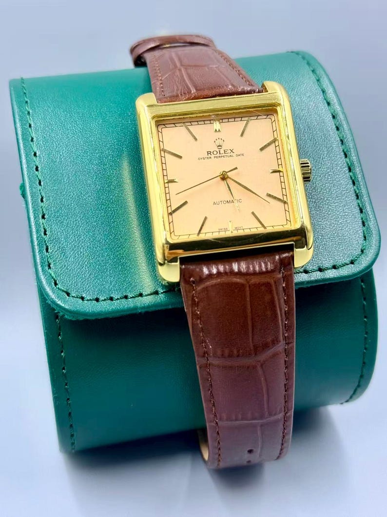 May include: A gold-toned Rolex Oyster Perpetual Date automatic watch with a rectangular face and brown leather band. The watch face is a light peach color with gold hands and markers. The watch is resting on a green leather case.