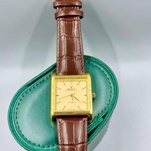 May include: A gold-toned rectangular Rolex watch with a brown leather strap. The watch face is a light gold color with black hands and text. The watch is displayed on a green leather case.