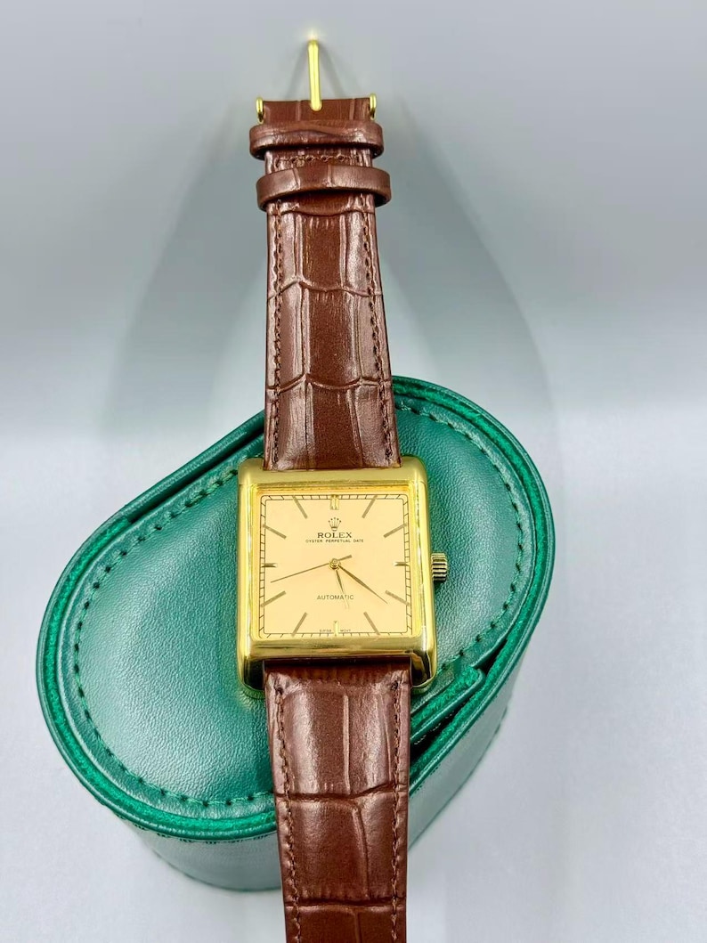 May include: A gold-toned rectangular Rolex watch with a brown leather strap. The watch face is a light gold color with black hands and text. The watch is displayed on a green leather case.