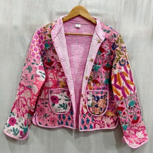 May include: A pink quilted jacket with a floral patchwork design. The jacket features a collared neckline, two front pockets, and wooden buttons. The interior lining is a lighter pink striped fabric.