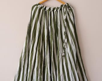 May include: A long, flowing skirt with vertical stripes in olive green and white. The skirt is hanging on a wooden hanger against a neutral background. The fabric appears lightweight and gathered at the waist.
