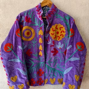 Puede incluir: Chaqueta morada con intrincados bordados florales en rojo, amarillo y verde. La chaqueta presenta un cuello alto y detalles en forma de corazón. Un ejemplo de artesanía vibrante.