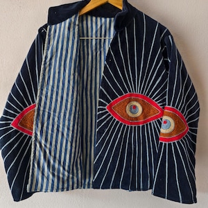 Puede incluir: Una chaqueta azul marino con un dise&ntilde;o &uacute;nico. La chaqueta presenta ojos bordados con detalles rojos y marrones en la parte delantera. L&iacute;neas blancas irradian de los ojos. El forro interior es a rayas azules y blancas.