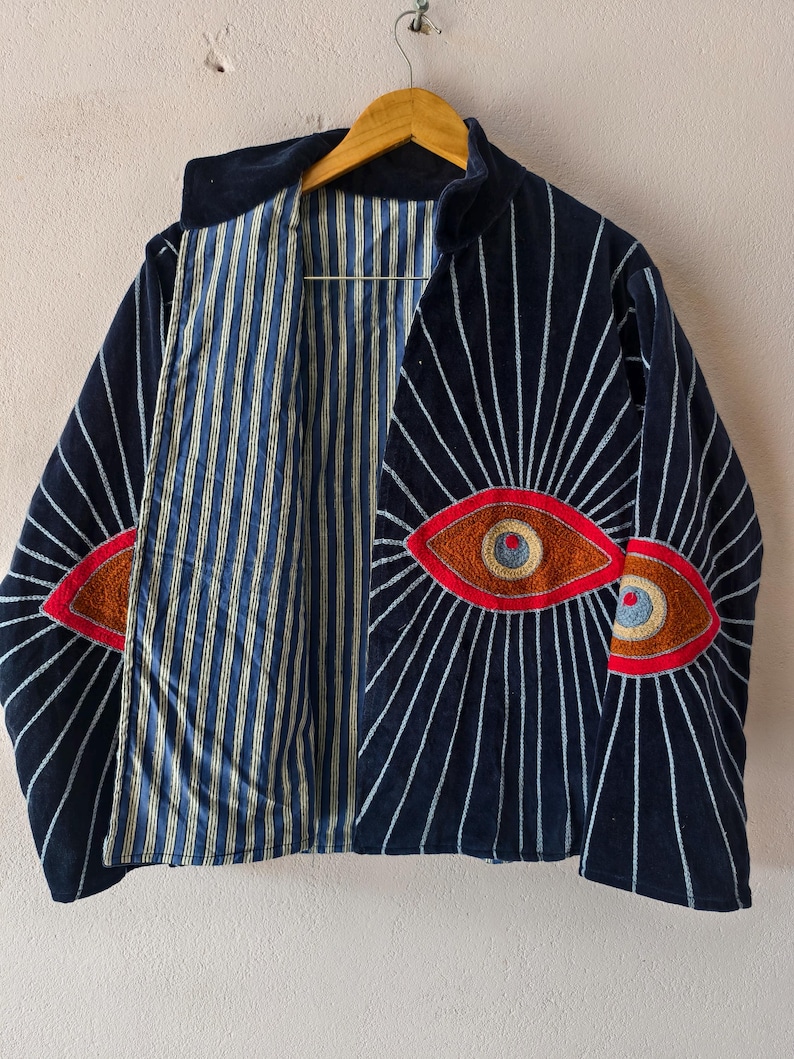 Puede incluir: Una chaqueta azul marino con un dise&ntilde;o &uacute;nico. La chaqueta presenta ojos bordados con detalles rojos y marrones en la parte delantera. L&iacute;neas blancas irradian de los ojos. El forro interior es a rayas azules y blancas.