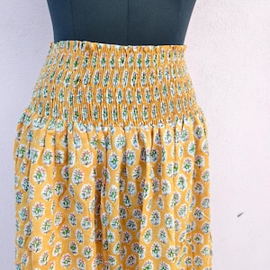 Puede incluir: Falda de color amarillo mostaza con estampado floral. La falda presenta una cintura fruncida y un diseño fluido. El estampado floral incluye detalles blancos, rosas y verdes. La falda se muestra en un maniquí.