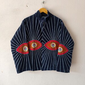 Puede incluir: Chaqueta azul marino con rayas verticales blancas que irradian desde los hombros. La chaqueta presenta dise&ntilde;os de ojos bordados en rojo, marr&oacute;n y dorado. La chaqueta est&aacute; colgada de una percha de madera.