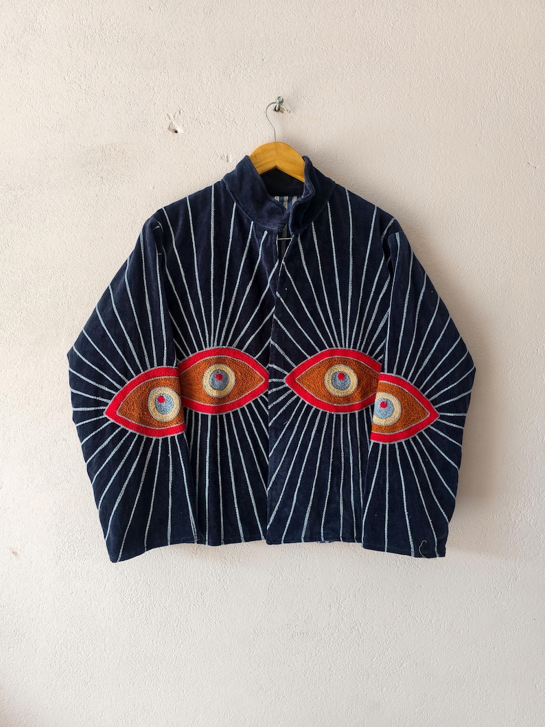 Puede incluir: Chaqueta azul marino con rayas verticales blancas que irradian desde los hombros. La chaqueta presenta dise&ntilde;os de ojos bordados en rojo, marr&oacute;n y dorado. La chaqueta est&aacute; colgada de una percha de madera.
