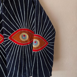 Puede incluir: Chaqueta azul marino con dise&ntilde;os de ojos bordados. La chaqueta presenta un patr&oacute;n de l&iacute;neas blancas que irradian de los ojos. Los ojos est&aacute;n detallados con bordados rojos, naranjas y azules. La chaqueta es de un material suave y aterciopelado.
