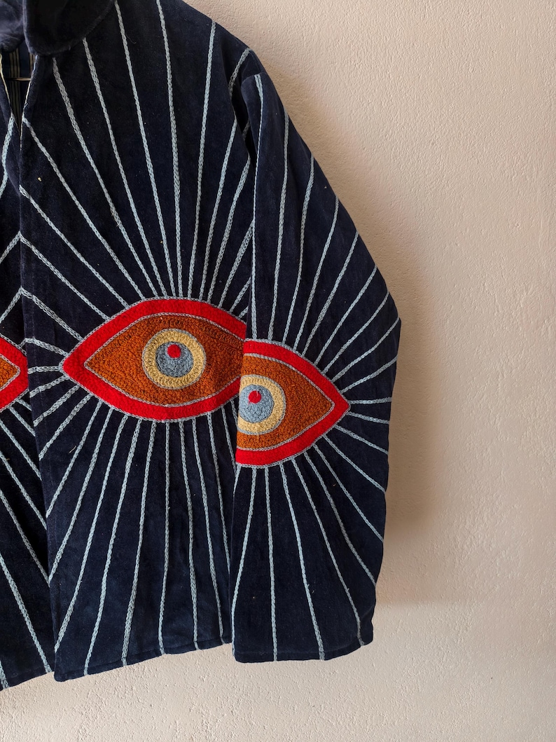 Puede incluir: Chaqueta azul marino con dise&ntilde;os de ojos bordados. La chaqueta presenta un patr&oacute;n de l&iacute;neas blancas que irradian de los ojos. Los ojos est&aacute;n detallados con bordados rojos, naranjas y azules. La chaqueta es de un material suave y aterciopelado.