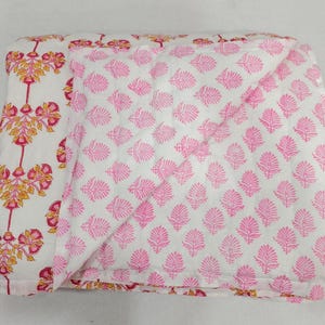 Könnte beinhalten: Eine weiße Steppdecke mit doppelseitigem Muster. Eine Seite zeigt ein sich wiederholendes rosa-gelbes Blumenmotiv mit vertikalen Stielen, während die umgeschlagene Ecke ein kontrastierendes Muster aus kleinen, sich wiederholenden rosa Blättern offenbart.