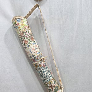 May include: A cylindrical yoga mat bag featuring a multi-colored floral block print pattern on a cream background. The bag has a long, thin, striped shoulder strap and a matching striped trim along the edges, with a tie closure at the top.