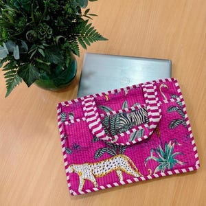 Pode incluir: Uma bolsa para laptop magenta com alça e acabamento listrados brancos e magenta. A bolsa apresenta um design de chita, zebra e plantas tropicais. Um laptop Dell e um arranjo floral verde estão no fundo.
