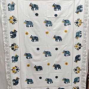 Könnte beinhalten: Weißes Tischtuch mit gestickten blauen Elefanten, gelben Blumen und dunkelblauen Akzenten. Der Rand hat ein sich wiederholendes Blumen- und Scroll-Design. Dekoratives Textil für die Heimdekoration.