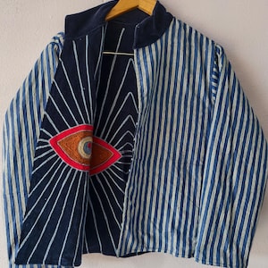 Puede incluir: Chaqueta a rayas azules y blancas con cuello y forro azul marino. La chaqueta presenta un dise&ntilde;o de ojo bordado en rojo, naranja y marr&oacute;n en el interior. La chaqueta est&aacute; colgada de una percha de madera.
