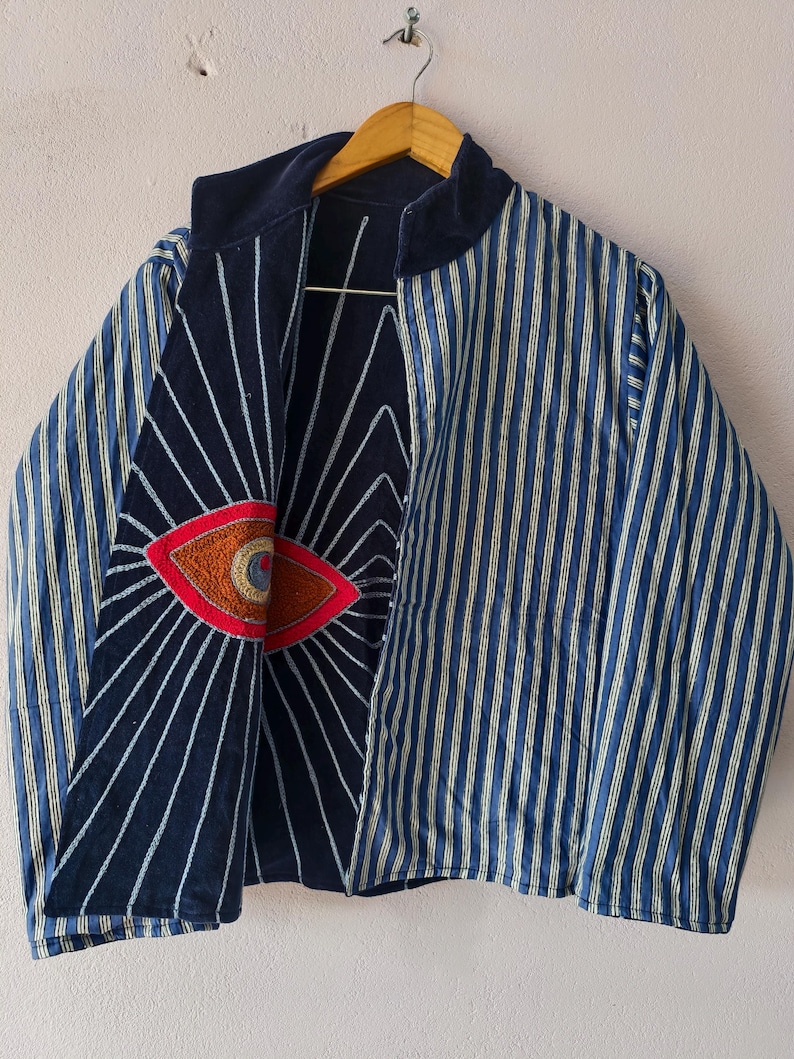 Puede incluir: Chaqueta a rayas azules y blancas con cuello y forro azul marino. La chaqueta presenta un dise&ntilde;o de ojo bordado en rojo, naranja y marr&oacute;n en el interior. La chaqueta est&aacute; colgada de una percha de madera.