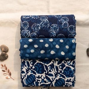 May include: Three folded pieces of fabric stacked vertically. The top piece is dark blue with white floral patterns. The middle piece is a medium blue with white polka dots. The bottom piece features a white background with dark blue floral motifs.
