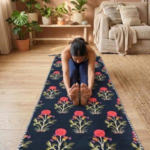 Pode incluir: Um tapete de ioga azul escuro com um padrão floral de flores vermelhas e folhas verdes. O tapete é retangular e tem uma borda branca. Uma pessoa se alonga no tapete.