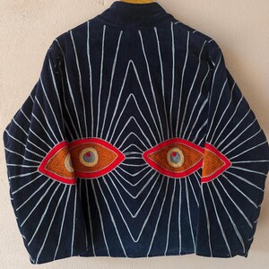 Puede incluir: Chaqueta azul marino con un dise&ntilde;o &uacute;nico. La parte trasera presenta dos dise&ntilde;os de ojos bordados en naranja y marr&oacute;n, rodeados de l&iacute;neas blancas radiantes. La chaqueta est&aacute; colgada en una percha de madera.