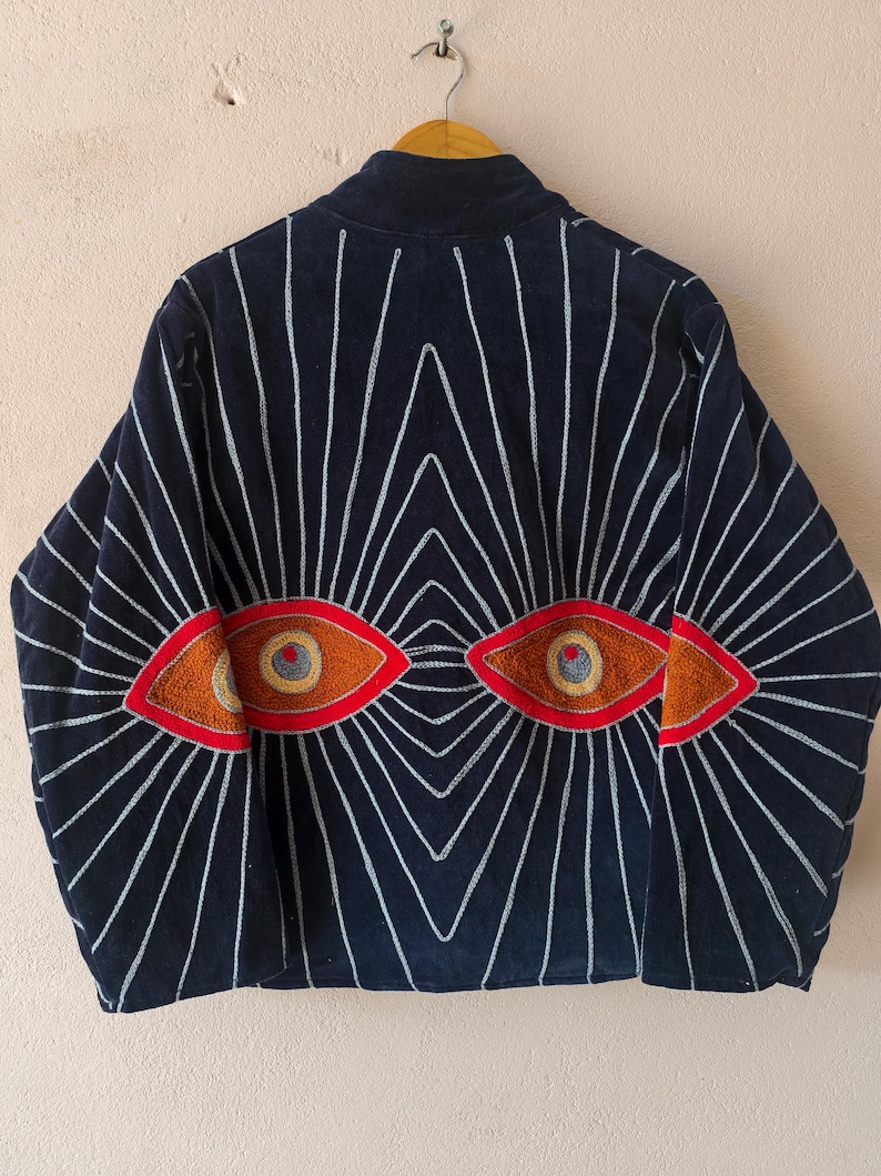 Puede incluir: Chaqueta azul marino con un dise&ntilde;o &uacute;nico. La parte trasera presenta dos dise&ntilde;os de ojos bordados en naranja y marr&oacute;n, rodeados de l&iacute;neas blancas radiantes. La chaqueta est&aacute; colgada en una percha de madera.