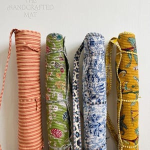 May include: Four yoga mat carriers in a row. The first is orange and white striped. The second is green with a floral pattern. The third is blue and white with a floral pattern. The fourth is yellow with a tropical design. The text "THE HANDCRAFTED MAT" is at the top.