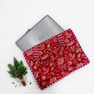May include: A red floral laptop sleeve with a silver laptop peeking out. The sleeve has a quilted design with a repeating pattern of pink flowers and green leaves. The laptop is a Dell brand. A small bundle of greenery is tied with twine.