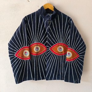 Puede incluir: Una chaqueta azul marino con dise&ntilde;os de ojos bordados con detalles rojos, marrones y azules. L&iacute;neas blancas irradian de los ojos. La chaqueta est&aacute; colgada en una percha de madera.