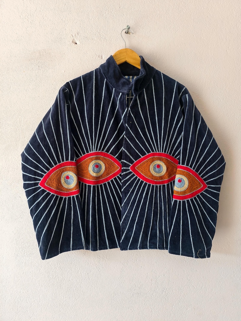 Puede incluir: Una chaqueta azul marino con dise&ntilde;os de ojos bordados con detalles rojos, marrones y azules. L&iacute;neas blancas irradian de los ojos. La chaqueta est&aacute; colgada en una percha de madera.