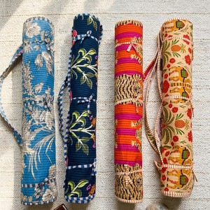 May include: Four colorful yoga mat carriers. The carriers are cylindrical and made of patterned fabric. One is blue with a floral design, another is navy with green and pink floral, the third is orange and pink striped, and the last is tan with a floral and elephant design.