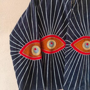 Puede incluir: Chaqueta azul marino con dise&ntilde;os de ojos bordados. La chaqueta tiene un cuello alto y l&iacute;neas blancas radiantes. Los ojos est&aacute;n delineados en rojo, con detalles marrones, amarillos y grises. La chaqueta es de un material suave y aterciopelado.
