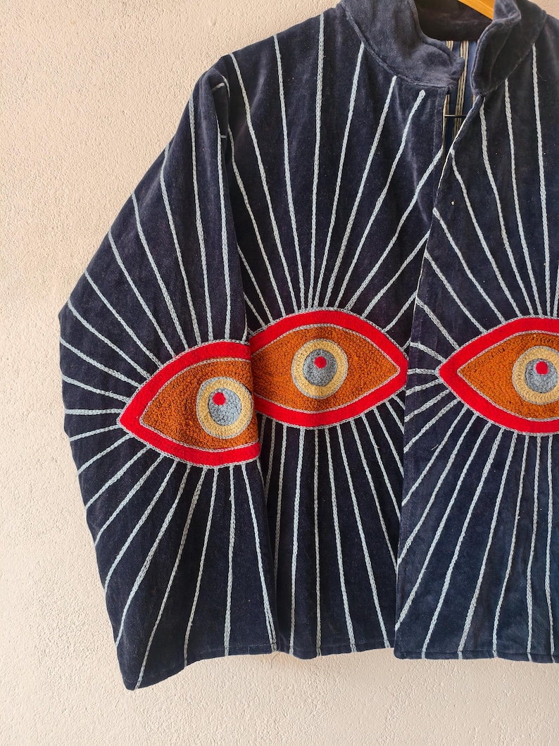 Puede incluir: Chaqueta azul marino con dise&ntilde;os de ojos bordados. La chaqueta tiene un cuello alto y l&iacute;neas blancas radiantes. Los ojos est&aacute;n delineados en rojo, con detalles marrones, amarillos y grises. La chaqueta es de un material suave y aterciopelado.