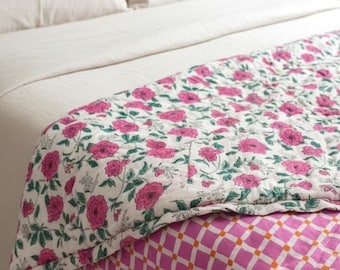 May include: A reversible quilt featuring a white base with a pink and green floral pattern on one side and a pink and white checkered pattern with small orange squares on the reverse side, draped over a bed with cream-coloured pillows and bedding.