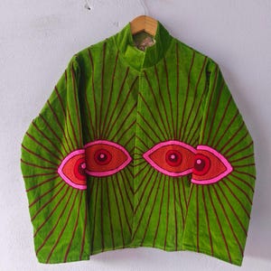 Puede incluir: Una vibrante chaqueta de terciopelo verde con cuello alto, adornada con diseños de ojos rojos y rosas bordados. La chaqueta presenta líneas radiantes en un tono más oscuro de rojo, creando un efecto visual llamativo. La chaqueta está colgada en una percha de madera.