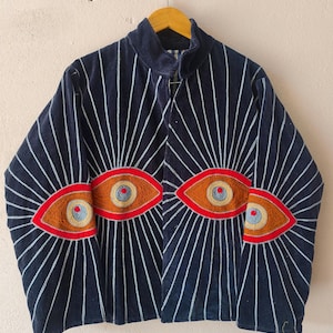 Puede incluir: Chaqueta azul marino con l&iacute;neas verticales blancas que irradian desde el centro. La chaqueta presenta tres dise&ntilde;os de ojos bordados en tonos naranja, rojo y azul. La chaqueta est&aacute; colgada en una percha de madera.